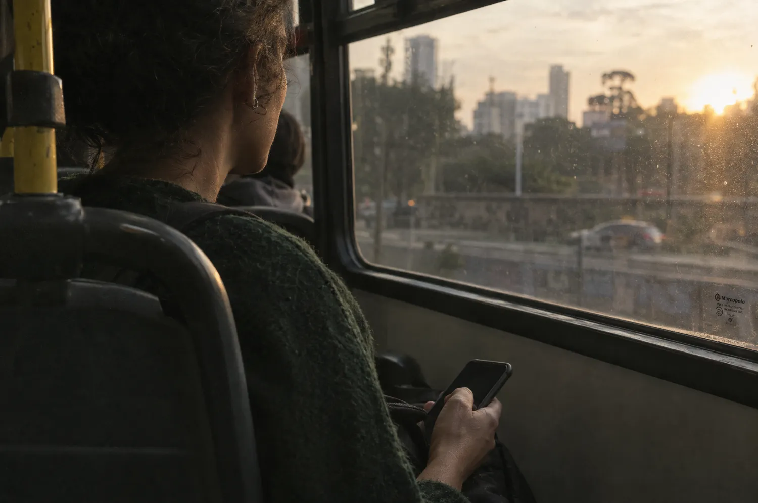 Passageira sentada junto à janela de um ônibus no fim da tarde, com o celular na mão.